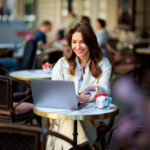 Cheerful woman sitting at a table, working on a laptop at an outdoor cafe. Attractive female wearing trench coat. Confident professional female having video call.