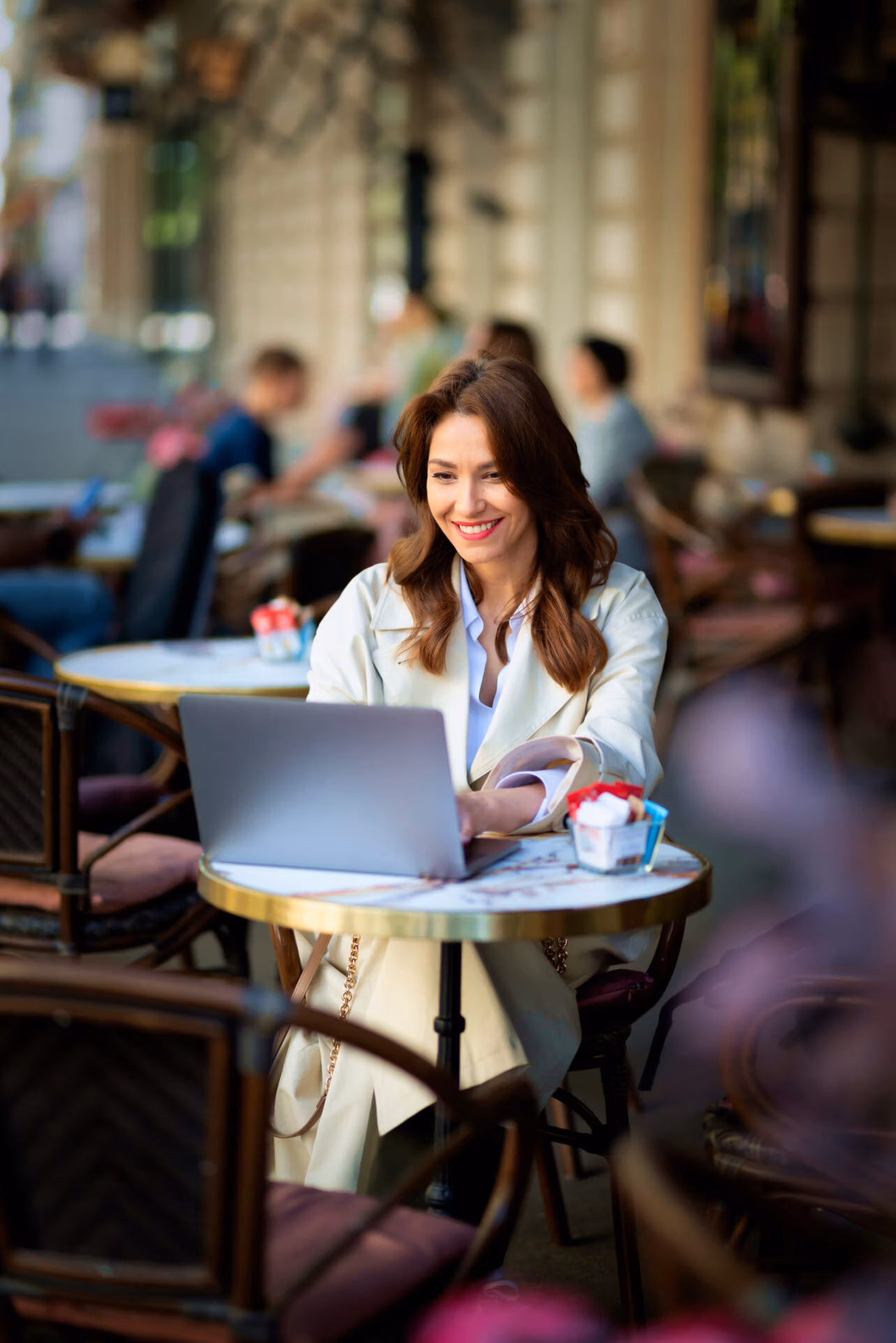 Cheerful woman sitting at a table, working on a laptop at an outdoor cafe. Attractive female wearing trench coat. Confident professional female having video call.